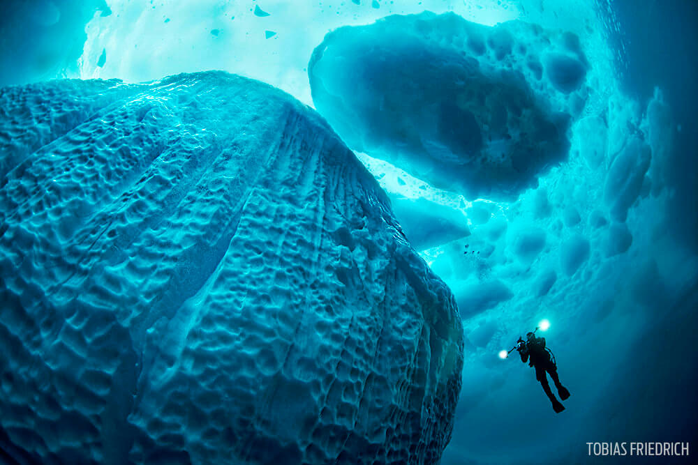 Diver swimming along an iceberg in crystal clear water holding a video camera with lights on, Tasiilaq Fjord, East-Greenland, Atlantic Ocean, Arctic, global warming.